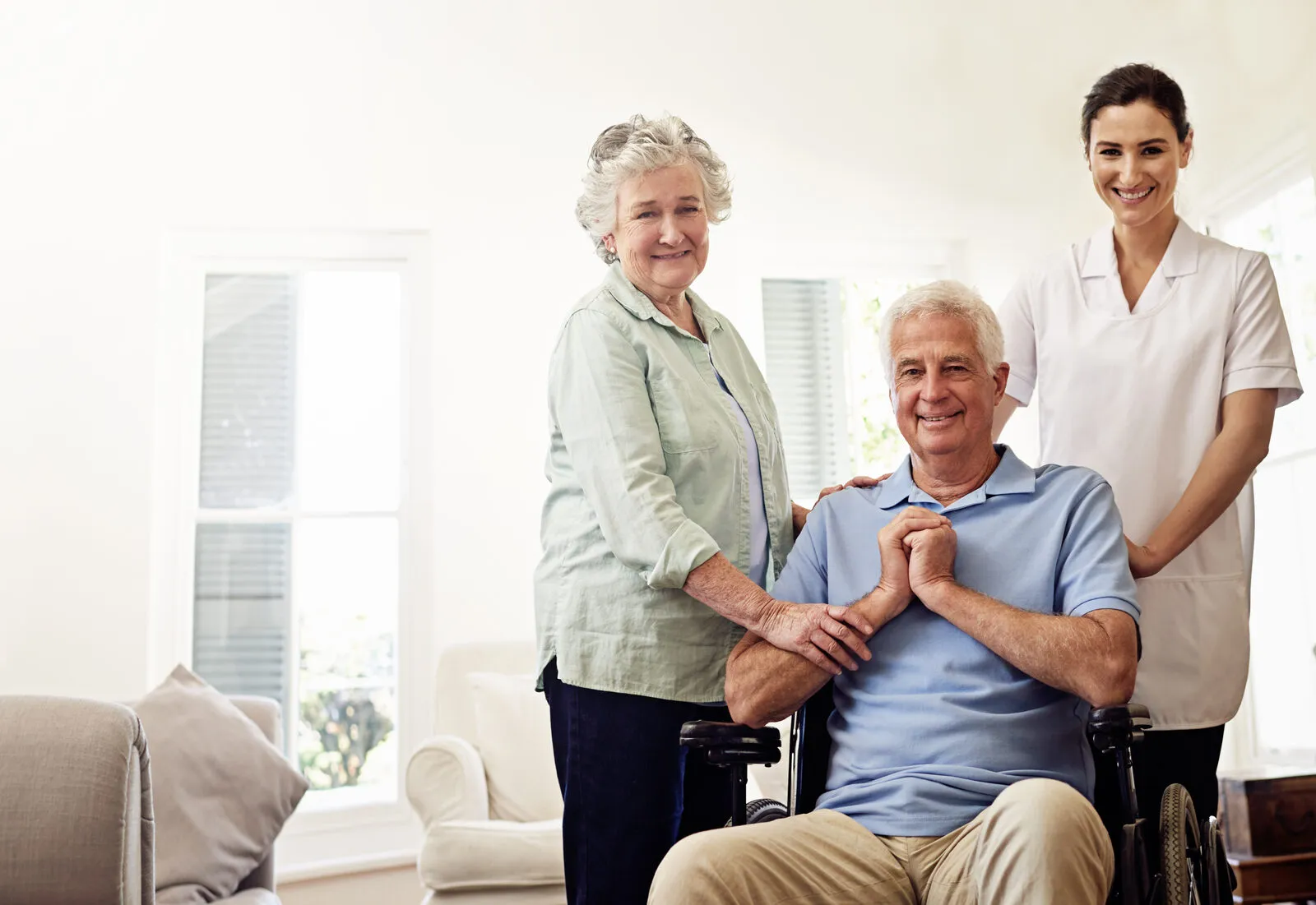 A SYNERGY HomeCare caregiver smiling with a senior couple in a bright living room, the man using a wheelchair