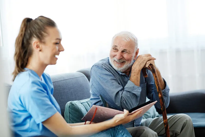 Home caregiver in scrubs smiling while chatting with happy senior man resting on cane at home