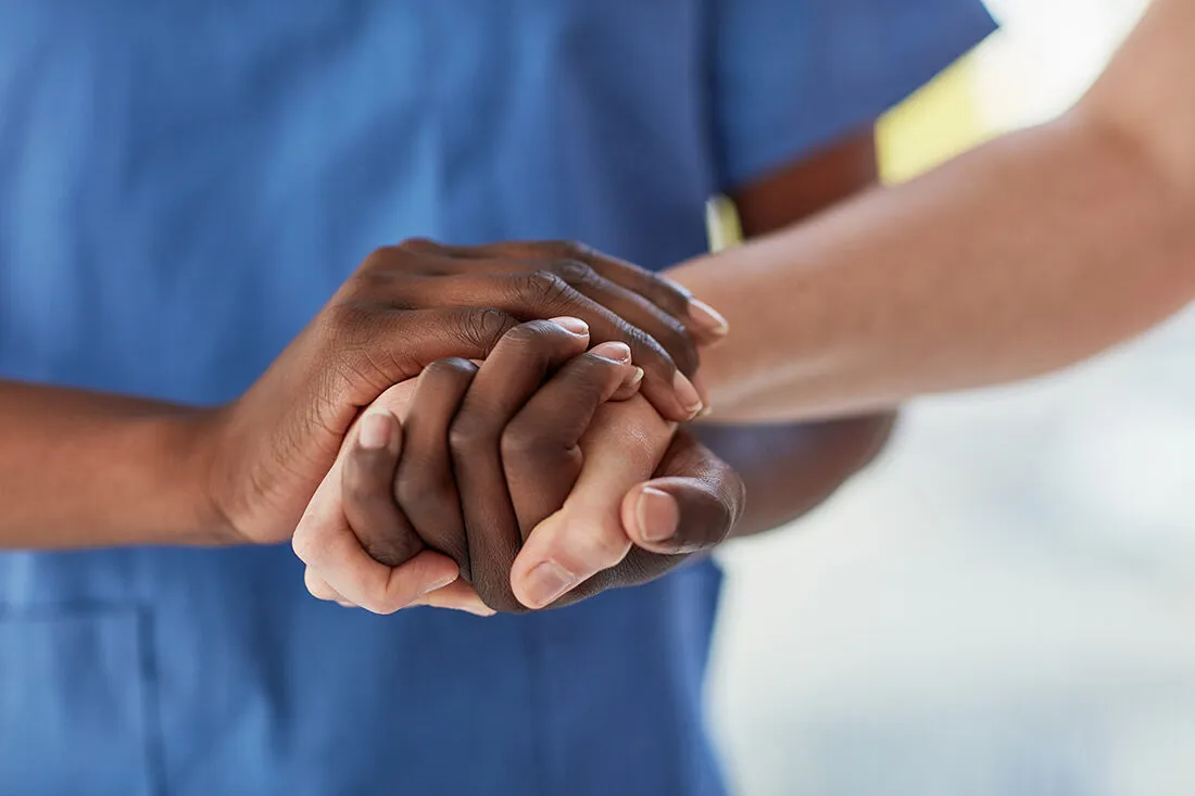 Black caregiver in blue scrubs holding the hands of an elderly white client: a moment of care