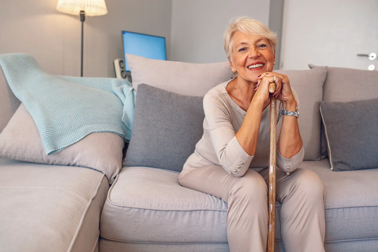 Active senior woman with short white hair sitting confidently on sofa, smiling with cane in hand