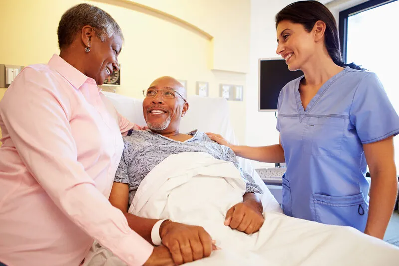 A smiling senior man recovering in a hospital bed with his wife and nurse at his side, beginning the transition to recovery at home